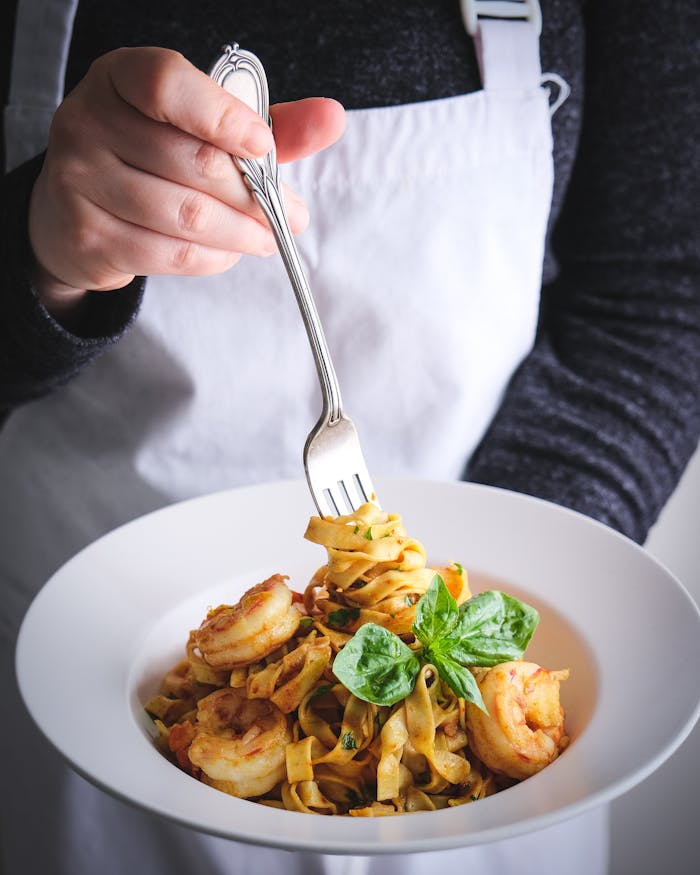 A close-up of shrimp pasta garnished with basil, served in a white dish and held by a person in a white apron.